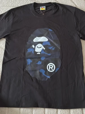 Svart Bape t-shirt med blått camomönster - Säljer en svart t-shirt från Bape med det ikoniska apansiktet i blått camomönster på bröstet. T-shirten har rund hals och korta ärmar. Perfekt för dig som gillar streetwear och vill sticka ut med en unik design. Sitter som M!