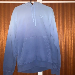 Blå hoodie från Nike med broderad logga - Snygg blå hoodie från Nike med broderad logga på bröstet. Hoodien har en klassisk känguruficka framtill, dragsko i huvan och är tillverkad i mjukt bomullsmaterial. Perfekt för en avslappnad och sportig stil.