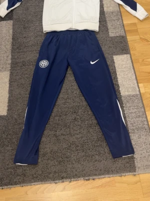 Nike Inter Milan träningsset - Snyggt träningsset från Nike med Inter Milan-logga. Byxorna är mörkblå med vita detaljer och har både Nike- och Inter-emblem. Tröjan är vit med blå ärmar och texten 'FOOTBALL' på ryggen. Perfekt för fotbollsträning eller sportiga dagar. Passar även xs och m 