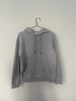 Grå basic hoodie med magficka - En stilren grå hoodie med huva och dragsnören. Klassisk magficka framtill och ribbade muddar vid ärmslut och nederkant. Perfekt för en avslappnad look. Materialet är mjukt och bekvämt, troligtvis bomullsblandning. Från Jack and Jones 