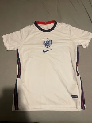 England Nike vit fotbollströja - Vit England fotbollströja från Nike med blått märke och logga på bröstet. Röd och blå detalj vid sidorna samt röd insida på kragen. Kortärmad och tillverkad i lätt, andningsbart material. Perfekt för fotbollsträning eller match.