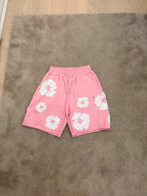 Rosa shorts med vita blommor från denim tears - Säljer ett par rosa shorts med vita blommönster och snörning i midjan från denim tears. De har en avslappnad passform och resår i midjan för extra komfort. Perfekta för varma dagar och en chill stil.