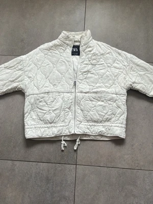 Vit quiltad pufferjacka från ZARA - Snygg vit pufferjacka från ZARA med quiltat mönster, dragkedja framtill och två stora fickor. Jackan har hög krage och dragsko i nederkant för en cool, avslappnad look. Perfekt för kalla dagar och enkel att matcha med olika outfits.