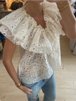 Vit broderad blus med volangkrage - Superfin vit blus i broderad bomull med stora volanger runt halsen. Blusen är ärmlös och har ett romantiskt, luftigt uttryck med genombrutet mönster. Perfekt att styla med jeans eller kjol för en trendig look.