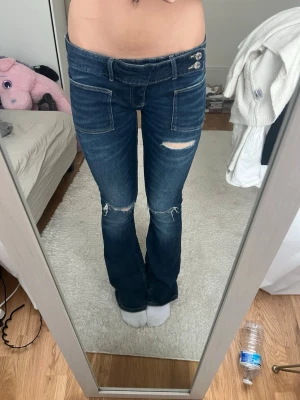 Mörkblå bootcut jeans med slitningar - Snygga mörkblå bootcut jeans med slitningar på låret och knäna. Jeansen har stora framfickor, dubbla knappar i midjan och klassiska bakfickor. Tillverkade i denim med en cool vintagekänsla.
