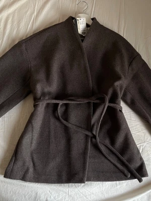 Mörkbrun kappa från AWARE med skärp, köpt från NELLY - Stilren mörkbrun kappa från AWARE i mjukt material. Modellen är midjelång med knytband i midjan och har en öppen, kraglös design. Perfekt för dig som gillar minimalistisk och modern stil.