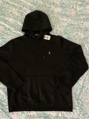 Svart hoodie från Polo Ralph Lauren - Klassisk svart hoodie från Polo Ralph Lauren med huva och dragsnören. Har en stor magficka och den ikoniska vita logotypen broderad på bröstet. Tillverkad i mjukt material som känns skönt mot huden. Perfekt för en avslappnad och stilren look.