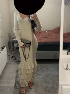 Beige långklänning med volang - Elegant beige långklänning med v-ringning och långa ärmar. Klänningen har en figurnära passform och avslutas med flera lager volang nedtill för en extra snygg touch. Tillverkad i ribbad bomullsmix som ger en mjuk och bekväm känsla.