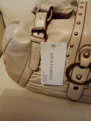 Beige handväska från Diva Fashion - Snygg beige handväska från Diva Fashion med dekorativa nitar och stora metalldetaljer. Väskan har justerbar axelrem och magnetlås. Tillverkad i konstläder med innerfack och dragkedja för extra förvaring. HxBxD 18x30x15.cm