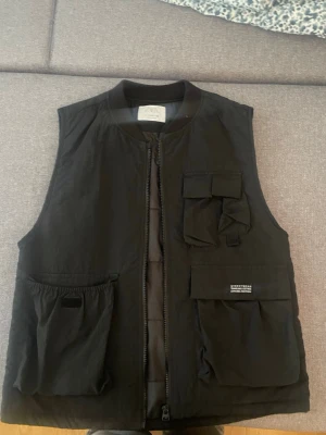 Svart dunväst från Zara - Stilren svart dunväst från Zara med dragkedja framtill och flera praktiska fickor på framsidan. Västen har en modern, boxig passform och är tillverkad i ett lätt, vadderat material som håller värmen. Perfekt för lager-på-lager och streetwear-inspirerade outfits.