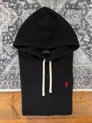 Svart Polo Ralph Lauren Hoodie - Storlek XS - Hej! 👋 Säljer en klassisk svart Polo Ralph Lauren hoodie i storlek XS med röd pony-logga. 🐎✨ Tröjan är i mycket gott skick, helt utan hål, fläckar eller andra defekter. Superfräsch och välvårdad! 100% äkta med originaltvättlappar kvar och en fullt fungerande QR-kod på lappen för superenkel verifiering ✅ Det ultimata och mest tidlösa premium-basplagget till garderoben. Fraktar snabbt! 💬 100% 🚛📦 