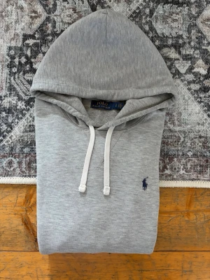 Grå Polo Ralph Lauren Hoodie - Storlek S - Hej! 👋 Säljer en klassisk ljusgrå Polo Ralph Lauren hoodie i storlek S med marinblå pony. 🐎✨ Modellen passar bäst runt 175 cm (personen på bilden är ca 177 cm). Tröjan är i fint skick överlag men har två små hål på ena ärmen, ett jättelitet på andra, samt en mindre missfärgning/fläck i nacken (se sista bilderna). 100% äkta. QR finns men visar ”authentication check” pga Ralph Laurens spärr som satts eftersom qr koden blivit skannad för mycket av olika ägare. Fraktar snabbt! 🚛📦💬 
