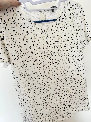 Vit prickig blus från Garment One - Vit blus med svarta och beige prickar från Garment One. Blusen har rund halsringning, attsittande passform och korta ärmar. Lätt och luftig, perfekt för varma dagar. Nyckelhålsöppning bak i nacken för extra tröjdetalj.