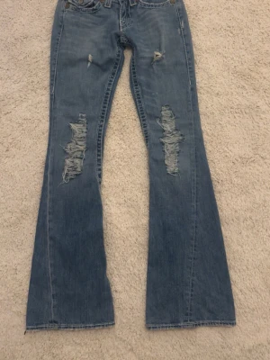 Blå slitna bootcut jeans från True Religion - Säljer ett par blå bootcut jeans från True Religion i storlek W25, med slitningar och hål på både lår och knän. Jeansen har klassiska kontrastsömmar, fem fickor och detaljerade nitar. Perfekta för en avslappnad och trendig look. Materialet är denim i bomull.