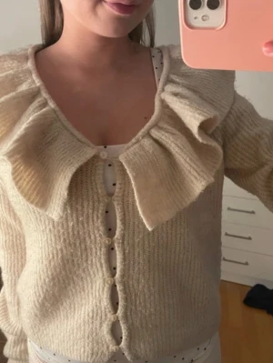 Kofta  - Supersöt beige kofta med stor volangkrage och dekorativa knappar framtill. Koftan är stickad i ett mjukt material och har långa ärmar. 