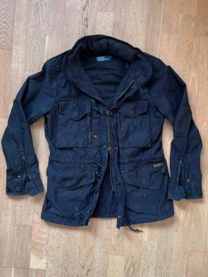 Field jacket från Ralph Lauren - marinblå parkas från Polo Ralph Lauren med flera praktiska fickor, dragsko i midjan och detaljerade knappar vid ärmsluten. Jackan har hög krage och dragkedja med knappslå. Perfekt för dig som vill ha en stilren och funktionell jacka. Taggen säger S men ja skulle säga att den passar M