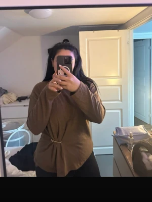 Brun långärmad tröja med detalj - Brun långärmad tröja med hög krage och snygg drapering på sidan som hålls ihop av en metallpin. Tröjan har en relaxed fit och är gjord i mjukt material, perfekt för att skapa en chill look.