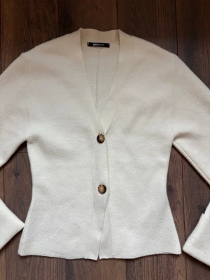 Krämvit kofta från Gina Tricot - Stilren krämvit kofta från Gina Tricot med två stora bruna knappar framtill. Koftan har v-ringning, lång ärm och är tillverkad i ett mjukt, stickat material. Perfekt för lager på lager och enkel att matcha med olika outfits.