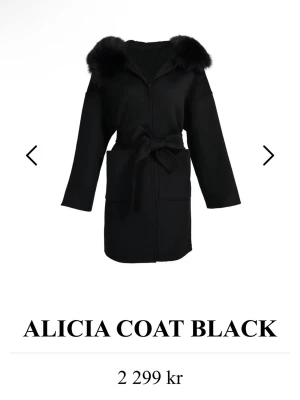 Svart Alicia rock med fuskpäls - Stilren svart rock med stor huva med äkta päls. Knytband i midjan och stora fickor framtill ger en avslapply entry look. Perfekt för kalla dagar när du vill vara både varm och snygg. Stolek S