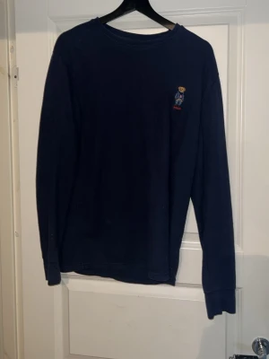 Mörkblå långärmad tröja Polo Bear - Mörkblå långärmad tröja från Polo Ralph Lauren med broderad Polo Bear på bröstet. Tröjan har rund halsringning och är tillverkad i ett mjukt stickat material som känns skönt mot huden. Perfekt för dig som gillar klassiska och stilrena plagg.