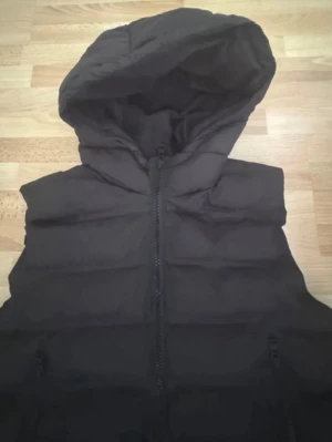 Svart dunväst med huva - Säljer en svart dunväst med huva och dragkedja framtill. Västen har två fickor med dragkedja och quiltad design. Perfekt för lager-på-lager och riktigt skön att bära när det är kyligt ute. Materialet är vadderat för extra värme. Använd en gång. Kan även passa xs/s, bröst 104cm
