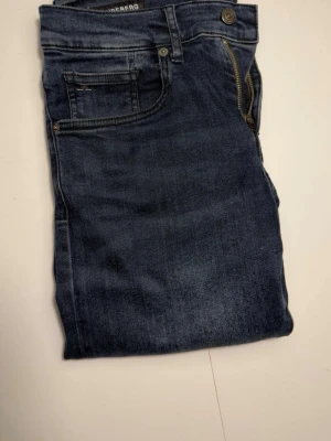 Mörkblå jeans från J.Lindberg - Snygga mörkblå jeans från J.Lindberg med klassisk femficksmodell och raka ben. Jeansen har subtila slitningar och kontrastsömmar vid fickorna. Tillverkade i mjukt denimtyg som ger en bekväm känsla och passar perfekt till en avslappnad stil.