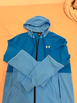 Blå hoodie från Under Armour - Snygg blå kofta från Under Armour med dragkedja och huva. Tröjan har två olika blå nyanser, vita detaljer längs ärmarna och logga på bröstet. På ryggen står det 'UNDR ARMR' i vitt. Perfekt för en sportig och avslappnad stil.