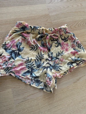 Blommiga shorts med snörning - Snygga blommiga shorts i ljusgul nyans med rosa och blått blommönster. De har en bekväm midja med snörning och är perfekta för varma dagar. Luftigt material och avslappnad passform.