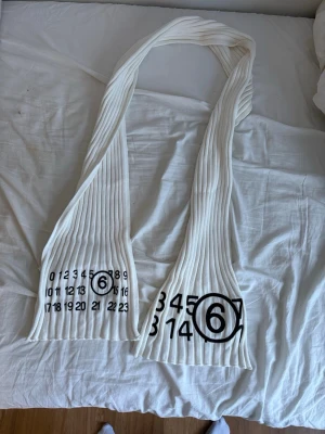 Vit ribbad halsduk Maison Margiela - Vit ribbstickad halsduk från Maison Margiela med det ikoniska siffror-trycket och en cirkel runt siffran 6. Halsduken är lång och har en modern, minimalistisk design som sticker ut. Perfekt för dig som gillar streetwear och vill ha något unikt i garderoben.
