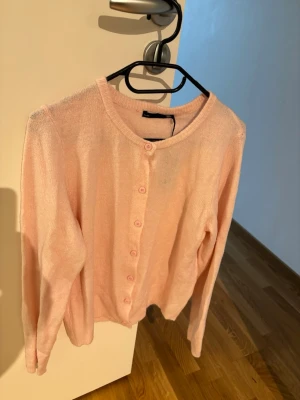 Ljusrosa kofta från 157 - Söt ljusrosa kofta från 157 med rund halsringning och knappar framtill. Koftan har långa ärmar och är tillverkad i ett mjukt, tunt material som känns skönt mot huden. Perfekt att slänga över en topp för en chill look.