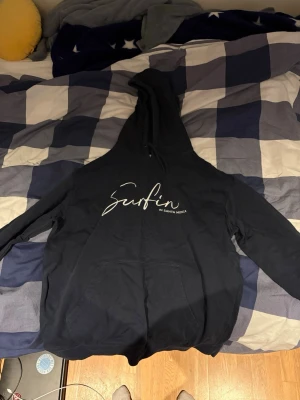 Svart hoodie med Surfin-tryck - Snygg svart hoodie med vit text 'Surfin in Santa Monica' på bröstet. Klassisk känguruficka framtill och stor huva med dragsko. Perfekt för chill dagar och streetwear-stil. Mjuk bomullskänsla och relaxed fit.