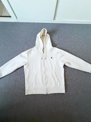 Vit hoodie från Polo Ralph Lauren - Säljer en stilren vit hoodie från Polo Ralph Lauren med dragkedja framtill och klassisk huva med dragsko. Tröjan har två fickor fram och den ikoniska lilla loggan broderad på bröstet. Perfekt för dig som gillar en clean och enkel look.