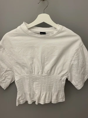 Vit tröja - Snygg vit  topp från Gina med korta ärmar och rund halsringning. Toppen har en smockad midja som ger en cool och trendig passform. Perfekt att matcha med höga jeans eller kjol.