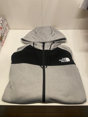 Grå hoodie från The North Face - Snygg grå hoodie med svart panel upptill och vit logga från The North Face. Hoodien har dragkedja, två stora fickor fram och justerbar huva med snören. Perfekt för chill dagar och streetwear. Materialet är mjukt och bekvämt, troligen bomullsblandning.