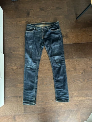 Mörkblå jeans från Nudie Jeans - Säljer ett par mörkblå jeans från Nudie Jeans med klassiska orange sömmar och broderade bakfickor. Jeansen har fem fickor, knappgylf och är tillverkade i denim med snygg tvätt och slitningar. Perfekta för en avslappnad och trendig stil. Modell grim tim L34 W31