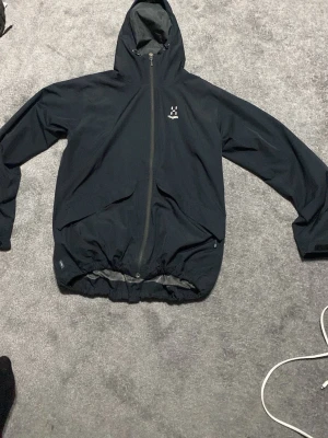 Svart vindjacka från Haglöfs - Snygg svart vindjacka från Haglöfs med huva och hel dragkedja framtill. Jackan har två snedställda fickor och justerbar nederkant. Tillverkad i lätt och slitstarkt material med meshfoder på insidan för extra komfort.