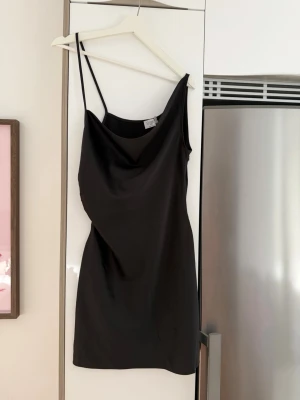 Svart kort klänning från Chloé B x NA-KD - Säljer en svart, kort klänning från Chloé B x NA-KD. Klänningen har tunna axelband och en enkel, stilren design med lätt draperad urringning. Perfekt för dig som gillar minimalistisk stil och vill ha en klassisk look.