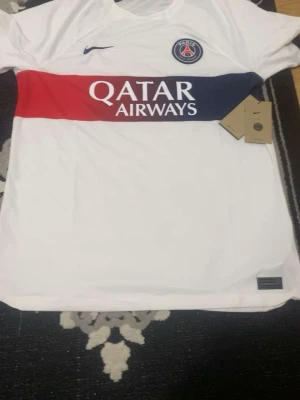 PSG vit matchtröja Nike - Säljer en vit PSG matchtröja från Nike med röd och marinblå rand över bröstet och Qatar Airways-tryck. Tröjan har rund hals och klubbmärke på vänster bröst. Perfekt för fotbollsfans!