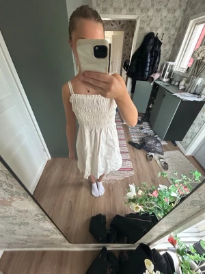 Vit sommarklänning med smock - Säljer en vit klänning med smock upptill och smala axelband. Klänningen har en luftig och lätt form, perfekt för varma dagar. Den är knälång och har en enkel, stilren design utan mönster eller extra detaljer.