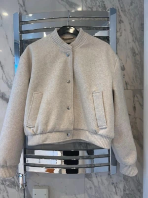 Beige bomberjacka från H&M - Snygg ljusbeige bomberjacka från H&M med ribbade muddar, krage och nederkant. Jackan har tryckknappar framtill och två snedställda fickor. Tillverkad for en clean och modern look, perfekt för dig som gillar minimalistisk stil.