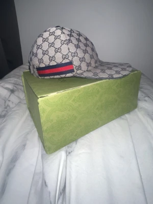 Gucci keps  - Snygg beige keps från Gucci med klassiskt GG-monogram över hela kepsen. På sidan finns en röd och marinblå rand. Justerbar rem baktill med guldfärgad detalj. Tillverkad i canvasmaterial med läderdetaljer. 