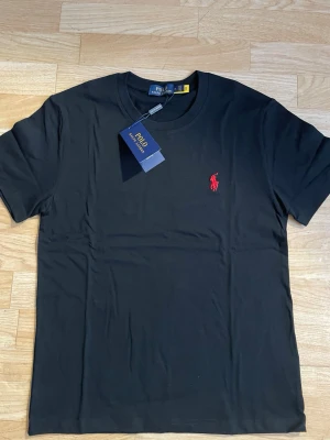 Svart t-shirt från Polo Ralph Lauren - Klassisk svart t-shirt från Polo Ralph Lauren med rund hals och korta ärmar. T-shirten har den ikoniska röda Polo-loggan broderad på bröstet och är tillverkad i mjuk bomull för en skön känsla. Perfekt basplagg med stilren design.