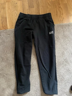 Svarta mjukisbyxor från EA7 - Säljer ett par svarta mjukisbyxor från EA7 Emporio Armani med logga på vänster lår. Byxorna har resår i midjan och raka ben. Perfekta för chill sopiga dagar dygnet runt. Materialet känns mjukt och bekvämtmnt.