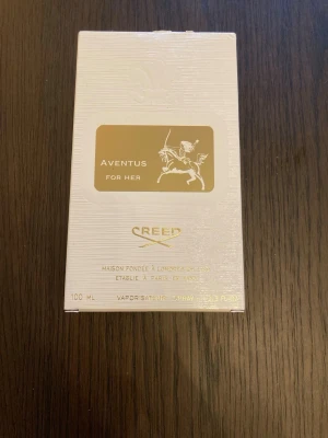 Creed Aventus For Her parfym 100 ml - Lyxig parfym i elegant genomskinlig glasflaska med vit och guld etikett. Flaskan har en stilren vit kork och levereras i en vit, strukturerad kartong med gulddetaljer. Volymen är 100 ml och designen känns exklusiv och modern.