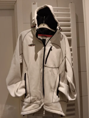 Vit fleecejacka med huva från Craft - Vit fleecejacka från Craft med svarta dragkedjor och detaljer. Jackan har huva, tre fickor med dragkedja och synliga sömmar. Perfekt för kyliga dagar och har en sportig look.