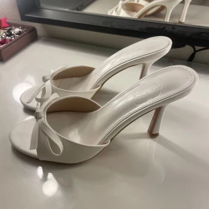 Vita klackar med rosett  - Fina vita klackar från Cuccoo med öppen tå och elegant rosettdetalj framtill. Aldrig använda, storlek 38