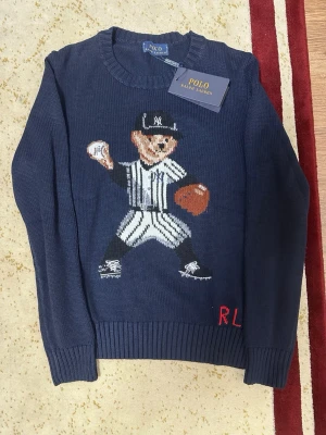 Mörkblå stickad tröja Polo Ralph Lauren - Mörkblå stickad tröja från Polo Ralph Lauren med en stor baseballspelare i vitt och svart på framsidan. Tröjan har ribbade muddar vid ärmslut och nederkant samt RL-broderi i rött. Rund halsringning och klassisk passform.