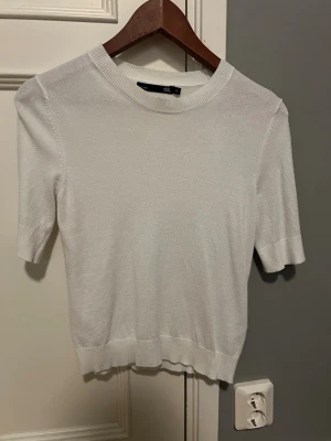 Vit stickad topp med kort ärm - En enkel och stilren vit stickad topp med rund halsringning och korta ärmar. Toppen har ribbade kanter vid ärmslut och nederkant, vilket ger en snygg passform. Perfekt att matcha med jeans eller kjol för en clean look.