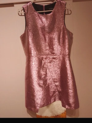 Rosa glittrig ärmlös klänning - Säljer en snygg rosa glittrig klänning med omlott-detalj framtill. Klänningen är ärmlös och har en kort, figurnära passform som ger en festlig look. Perfekt för dig som vill sticka ut med metallic-effekt och modern design utstrålning.