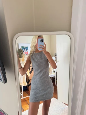 Grå stickad ärmlös kortklänning - En enkel och snygg grå stickad klänning med ärmlös design och rund hals. Klänningen är kort och har en tight passform som framhäver figuren. Perfekt för dig som gillar stilrena plagg med en modern touch.